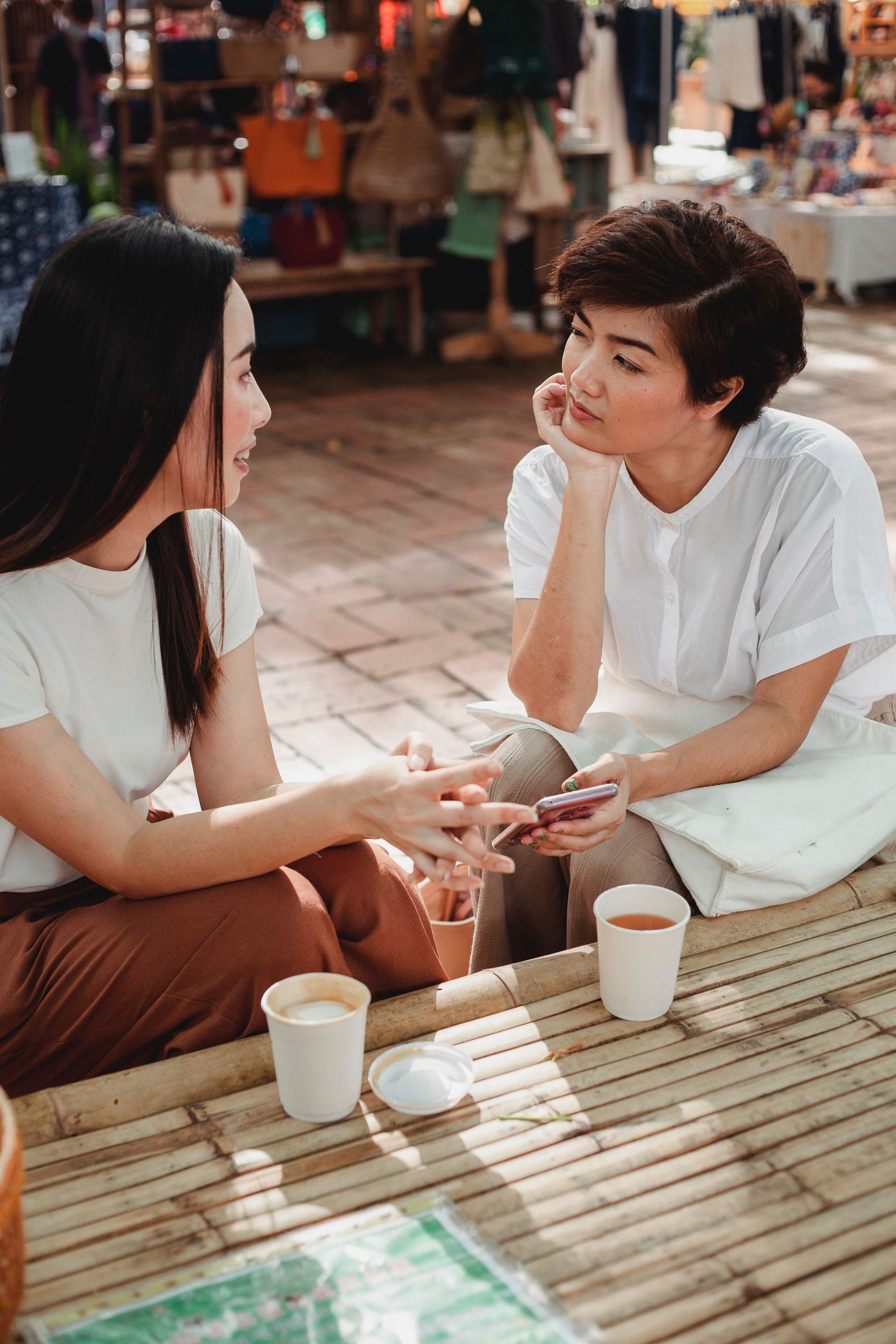 Two women sitting at an outdoor coffee shop. One of them is holding a phone.