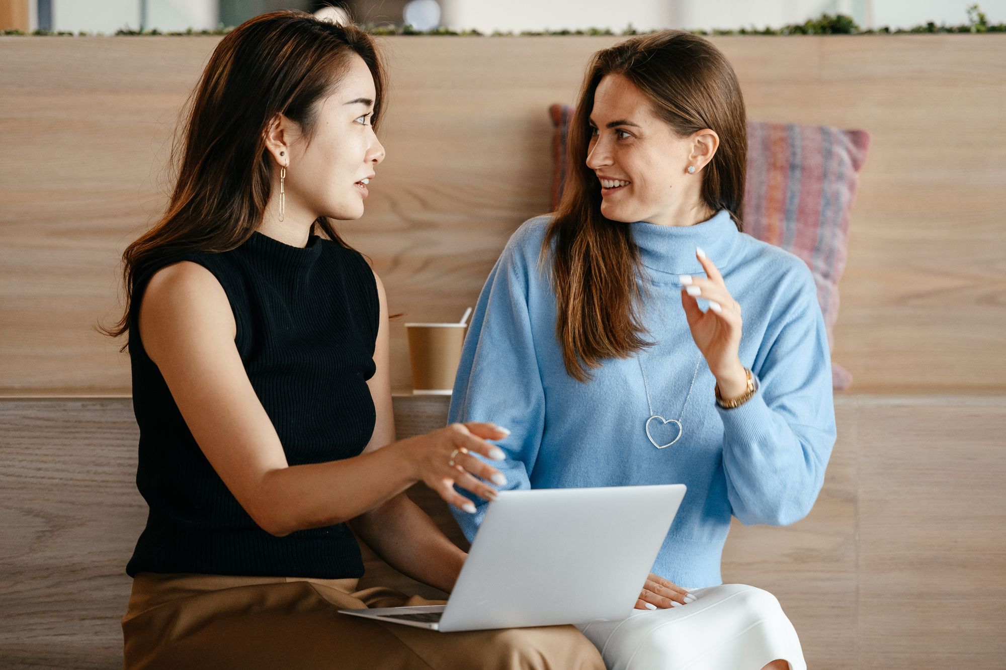 Two women talking to each other about work with a laptop in front of wooden stadium seating