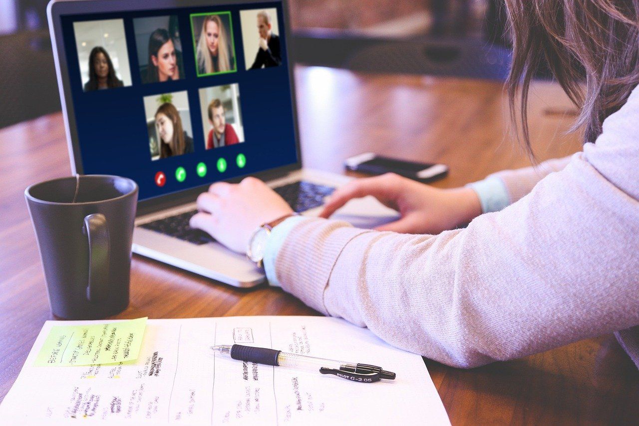 A laptop open with six faces visible for a video call. The laptop is next to a cup of tea and an open notebook. Someone is typing on the open laptop.