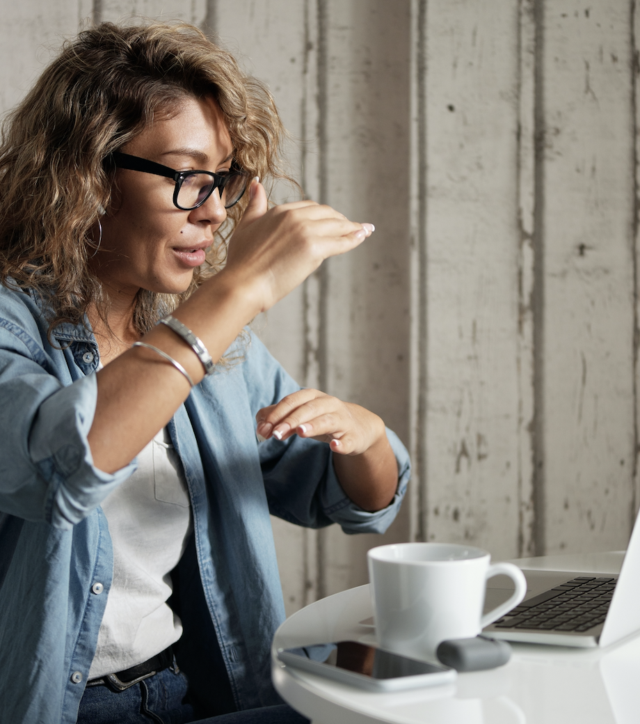 A woman in a denim shirt talking on a laptop with a cup of coffee and a smart phone