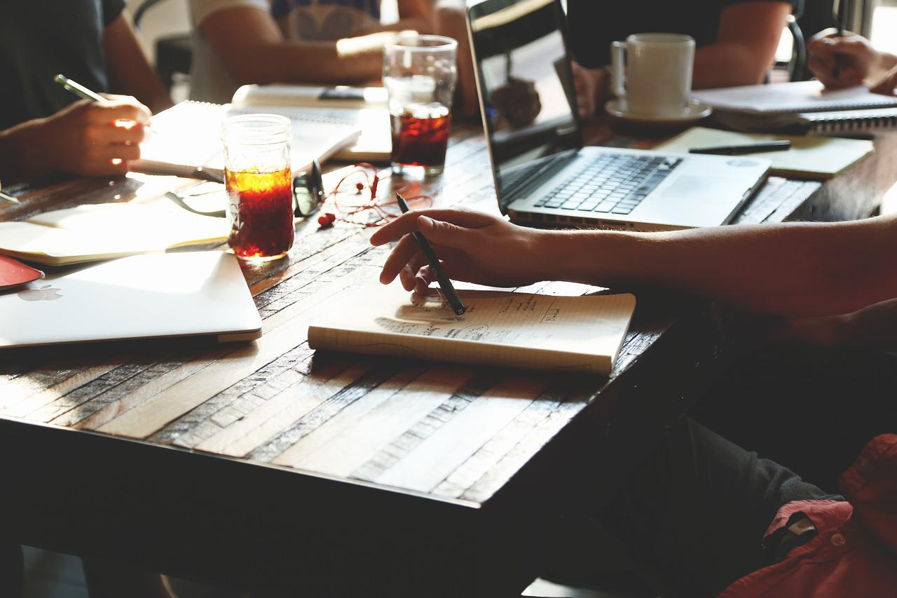 A photo of a group of people sitting around a table with laptops, notebooks, and coffee