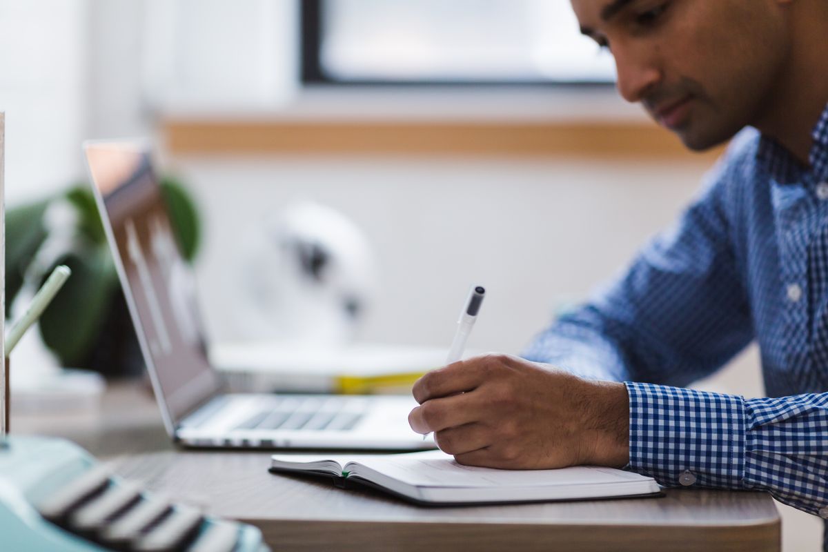 Photograph of a man writing in a notebook next to a laptop