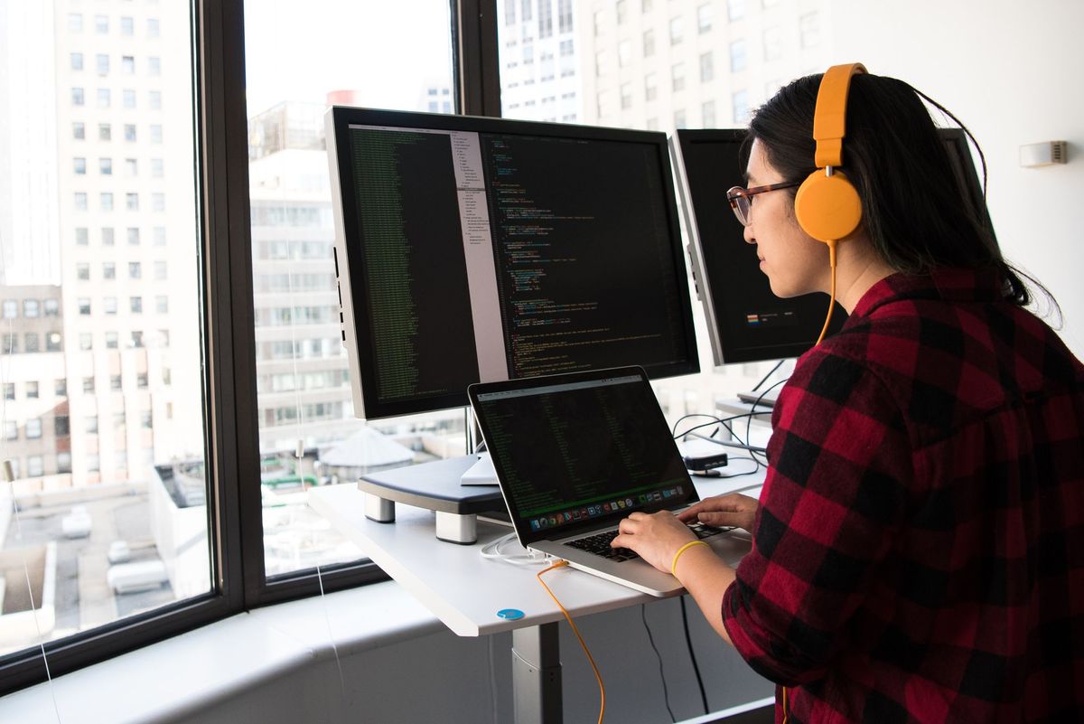 Woman wearing headphones writing code in front of a laptop and a large monitor